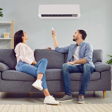 A couple relaxing on a couch, using a remote to control an air conditioner.