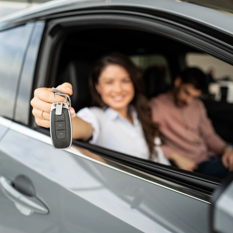 Smiling woman holding up car keys, celebrating purchasing a new vehicle. Man sitting next to her inside the car