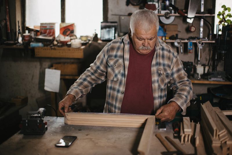 Senior man focusing on measuring a wooden frame using a tape measure in his workshop