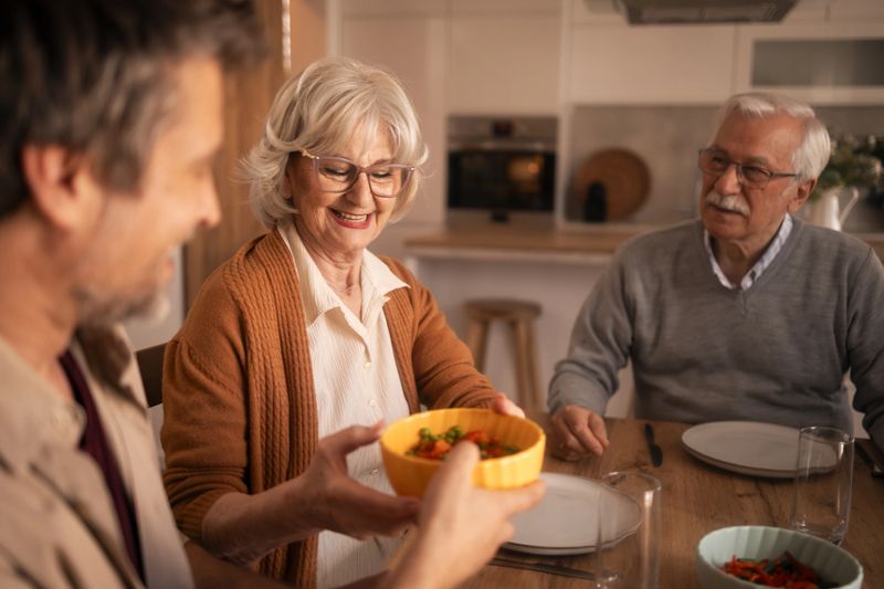 Family members enjoying a happy lunch together at home, with an adult son passing a bowl of fresh salad to his smiling senior mother at the dining table