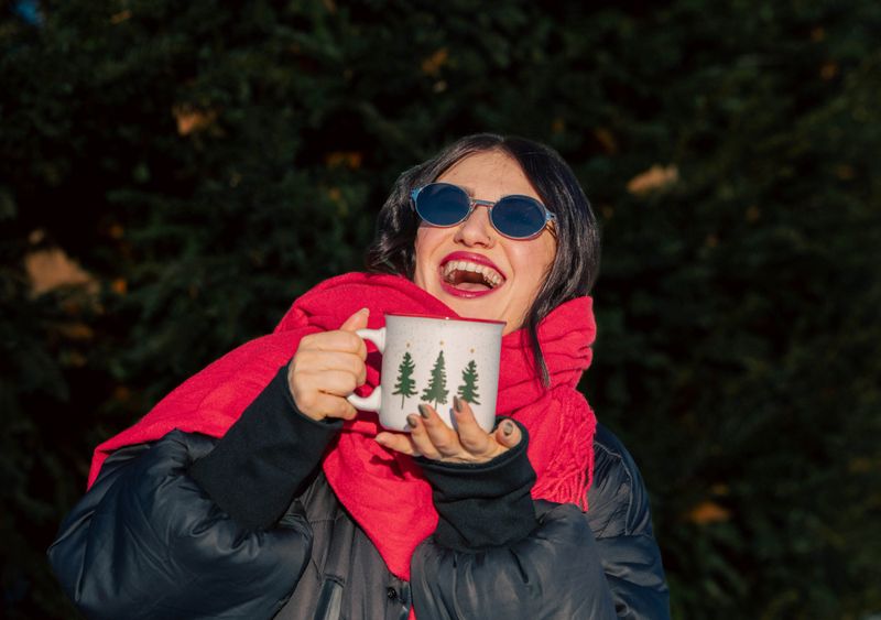 Female with dark hair, wearing sunglasses and a red scarf, joyfully holds a mug with tree design, surrounded by greenery, embodying a festive winter atmosphere