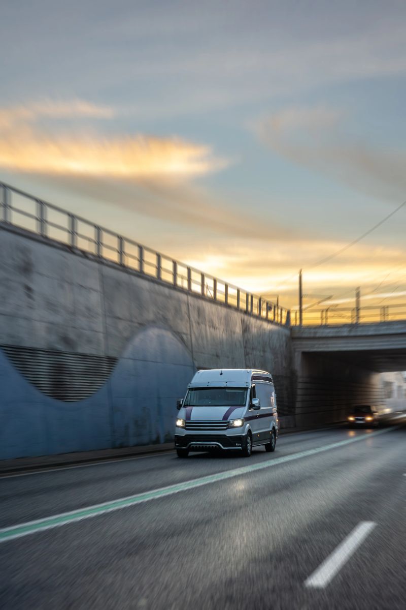 A white van drives beneath an underpass where concrete walls and shadows shape a strong urban mood. Warm light on the horizon adds cinematic contrast for delivery and transport themes.