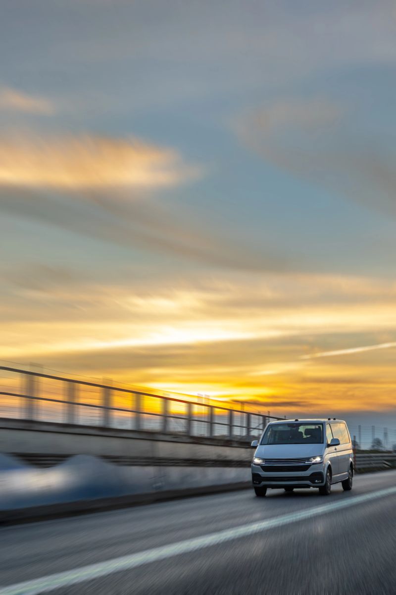 A light van drives along the highway beside bridge rails with motion blur that highlights speed and flow. Warm horizon light adds a cinematic mood for delivery and mobility concepts.