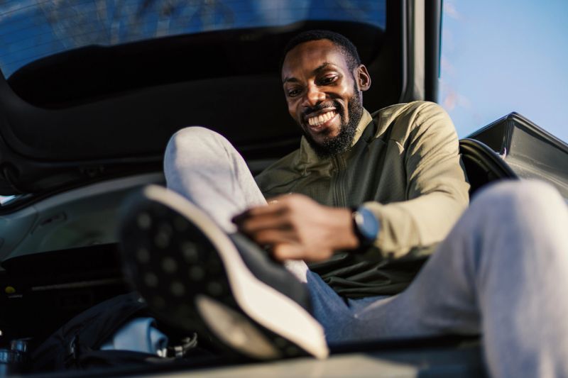 Happy young adult black man wearing activewear sitting in car trunk smiling while lacing up his running shoes, preparing for an outdoor fitness journey