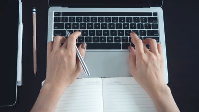 Hands typing on a laptop keyboard with an open notebook.