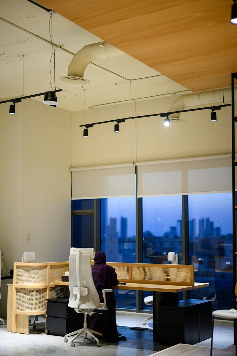 Full length shot of a Saudi businesswoman in an abaya working at a desk inside a modern office with an exposed ceiling, wooden finishes and city skyline visible through large windows at night.