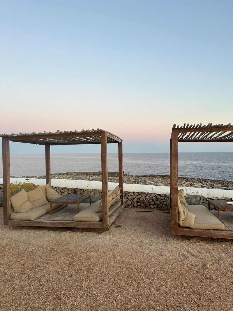 Cozy wooden pergola with soft cushions by the sea at sunset. Minimal coastal lounge area with natural textures, calm atmosphere, slow living, travel and seaside relaxation concept.