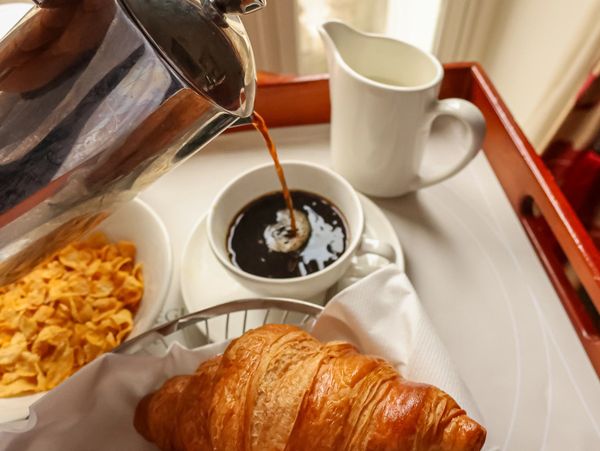 Coffee being poured with croissant and cornflakes on a breakfast tray.