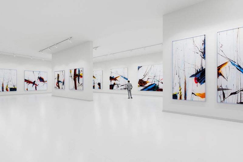 The interior of a modern and minimalist art gallery. The floors and walls are white, with large abstract expressionist paintings in black, blue, yellow, and red hanging on the white walls. A man is looking at the paintings.