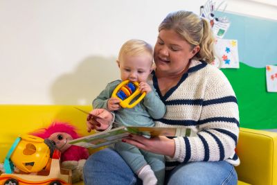 A woman reading a book to a baby holding a toy in a colorful playroom.