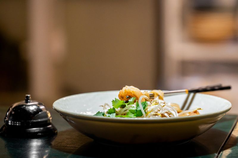 A close-up, shallow depth of field shot of Thai Pad Thai noodles with shrimp, bean sprouts, and cilantro in a white bowl at a restaurant table