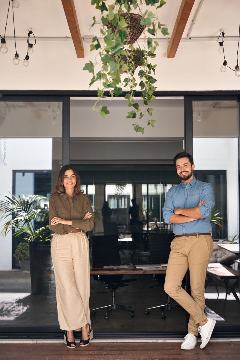 Two business partners executives standing looking at camera. 2 managers, company professionals oe entrepreneurs posing for corporate vertical portrait in modern cozy office with green plants.