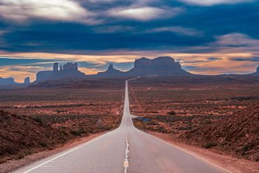 Long empty road leading to desert mesas under dramatic sky.