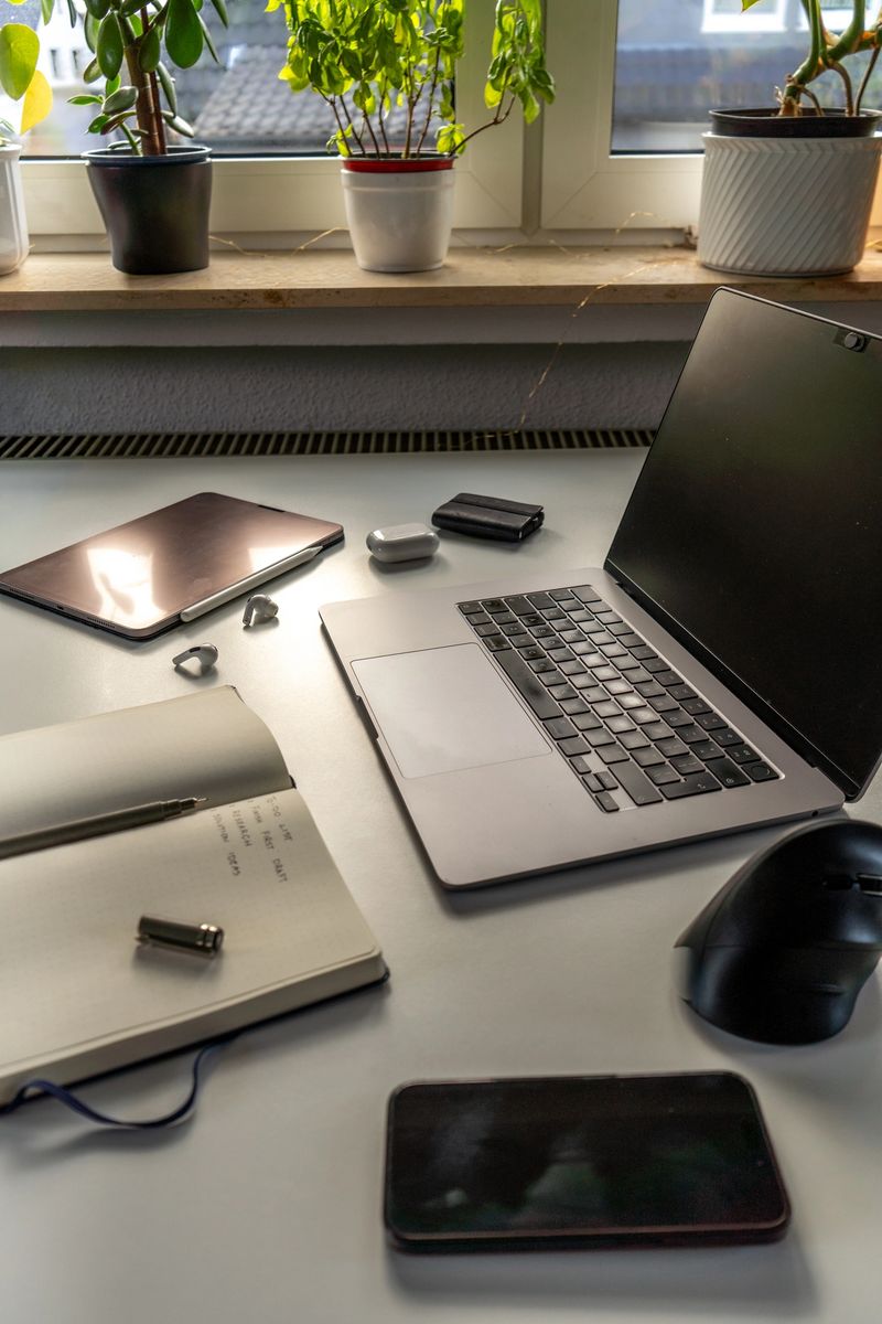 Modern home office desk with laptop, notebook, tablet, smartphone, wireless mouse, and earphones near a window with green houseplants, remote work concept.