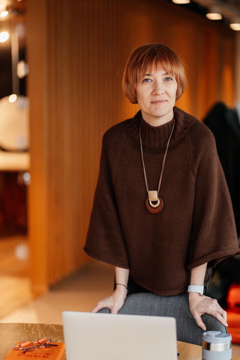 Confident professional woman in brown knit sweater and statement necklace, standing at desk with open laptop in warm modern interior. Experienced specialist, leadership presence. Vertical portrait.