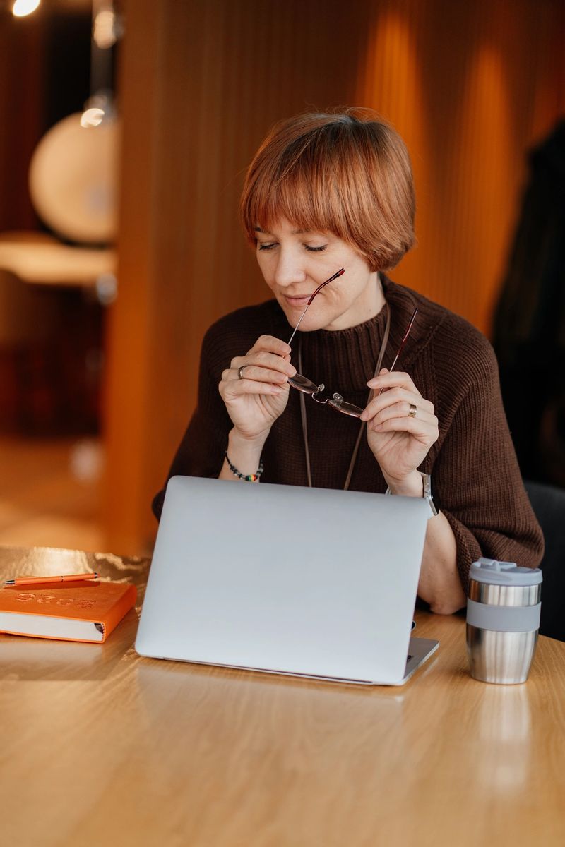 Analytical woman in her 40s with short hair wearing brown knit sweater, holding eyeglasses while looking at laptop at wooden table. Expert work process, calm professional mood,