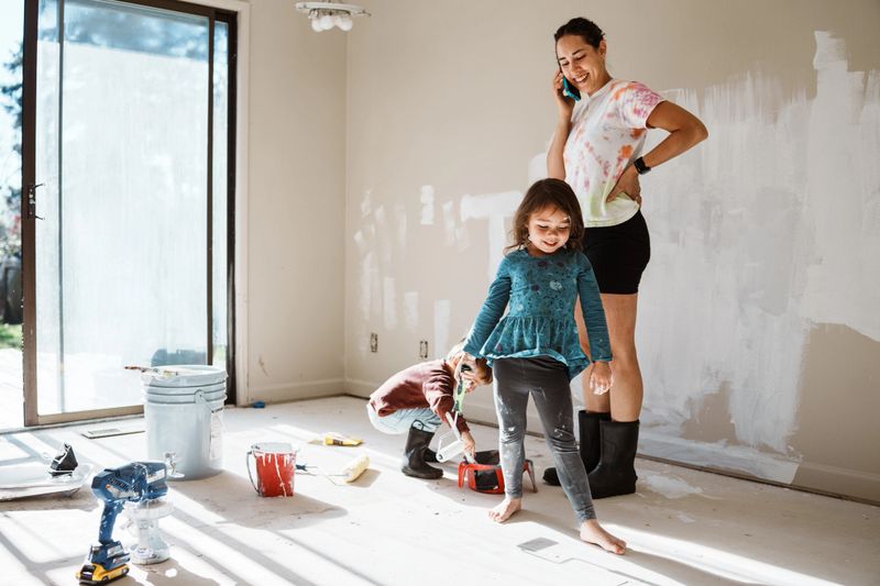 A Eurasian woman smiles during a phone call as her two young children paint and play in a bedroom under renovation.