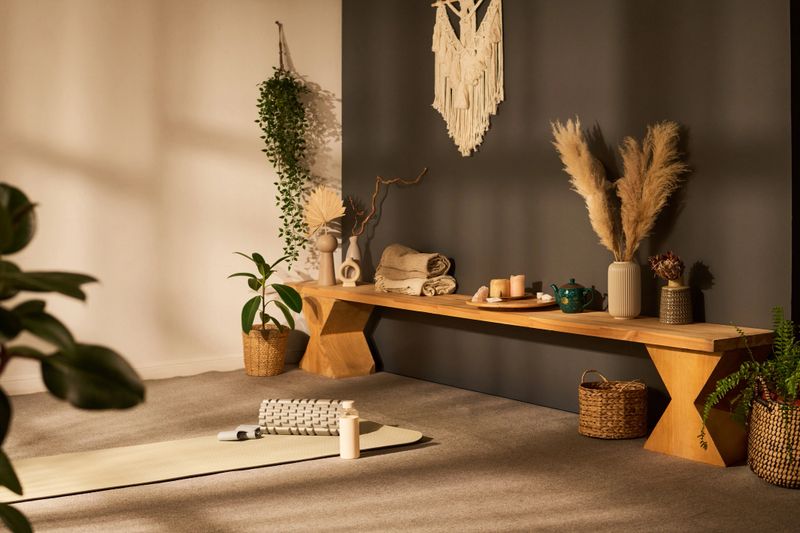 Yoga studio interior featuring wooden bench with decorative vases, candles, pampas grass, woven baskets, yoga mat, foam roller, water bottle, plants arranged on carpeted floor