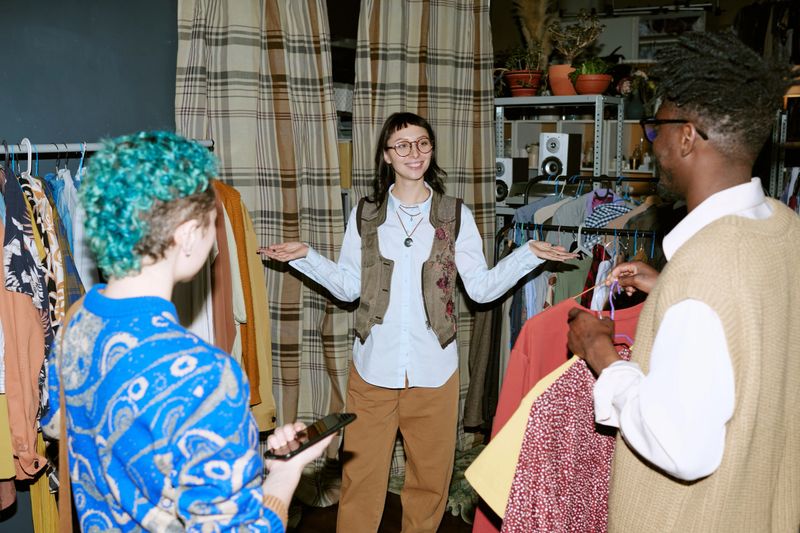 Young adult biracial woman standing with arms outstretched smiling while talking to young adult Black man and young adult Caucasian woman, browsing second hand clothing store racks