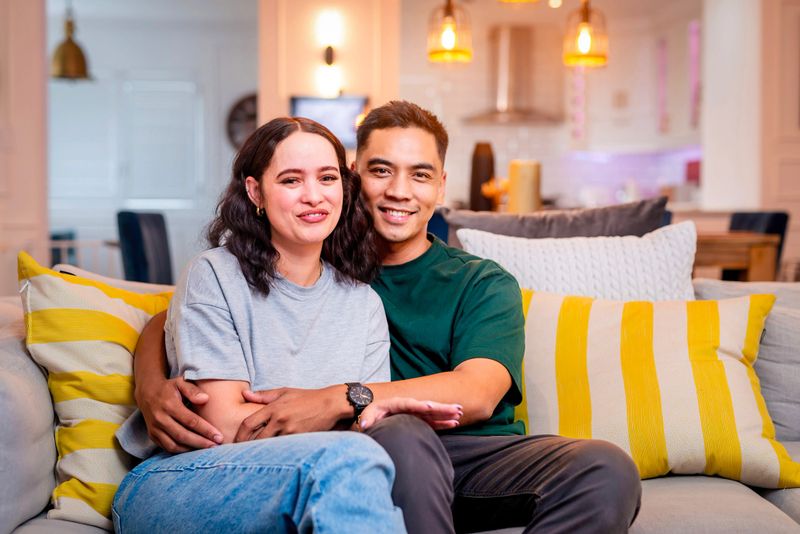A smiling couple sits close on a sofa in a bright, modern living room. Warm lighting, yellow striped cushions, and relaxed body language convey comfort, connection, and everyday joy.