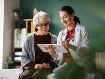An older woman and a doctor sitting next to each other looking at a tablet the doctor is holding.