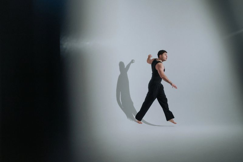 male soloist walking across spotlighted floor, cinematic silhouette and long shadow create moody vignette, wide studio composition