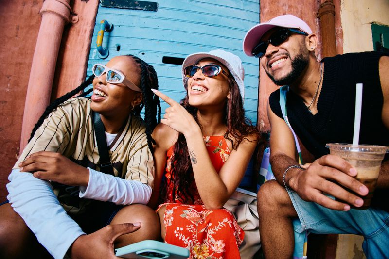Smiling group of young friends pointing and laughing while sitting outdoors beside a vibrant building, expressing curiosity and excitement during casual city exploration.