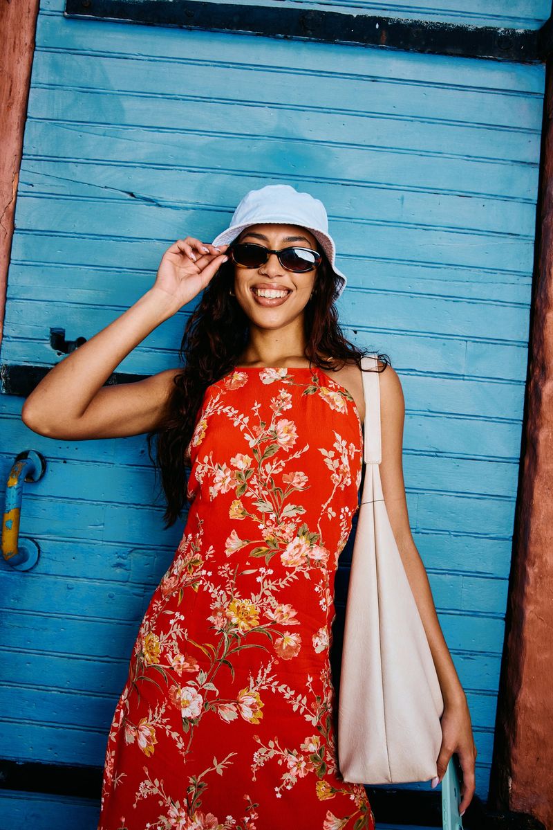Confident young woman in her 20s standing against a blue wall, wearing a colourful floral sundress, sunglasses, and hat while enjoying a sunny urban moment.