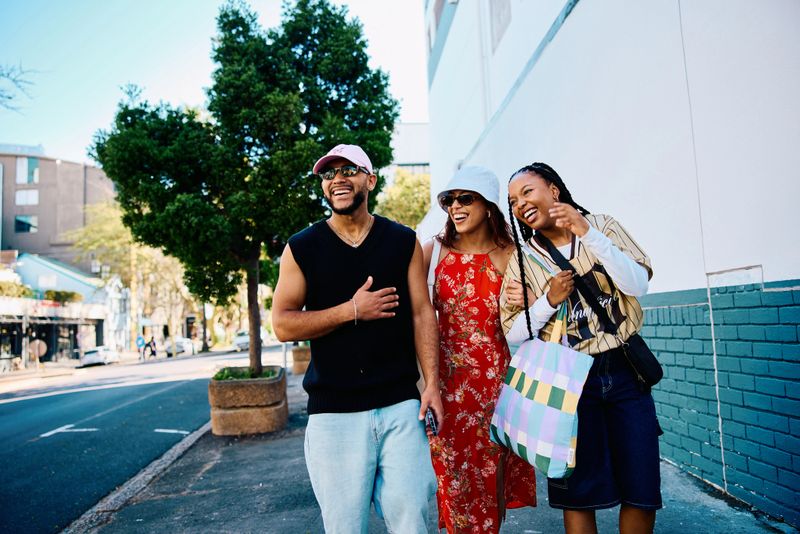 Vibrant young adults in their 20s walking arm in arm along a sunlit city street, sharing conversation and laughter during a carefree urban travel experience.