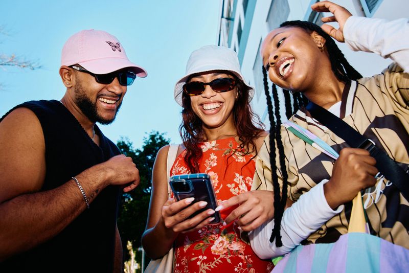 Cheerful young adults in their 20s standing on a bright city street, laughing while looking at a smartphone used for navigation during summer sightseeing.