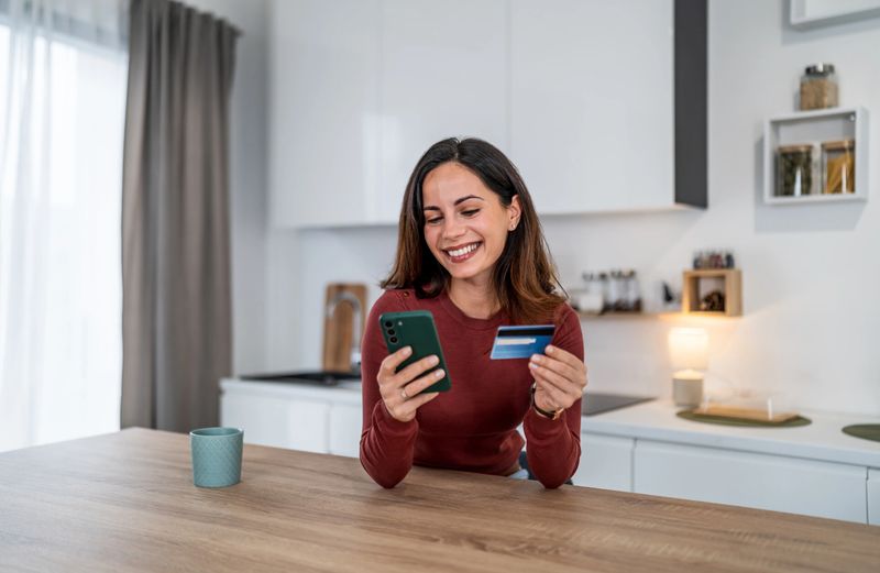 Happy young woman making online payment with smartphone and credit card, enjoying retail therapy from her kitchen