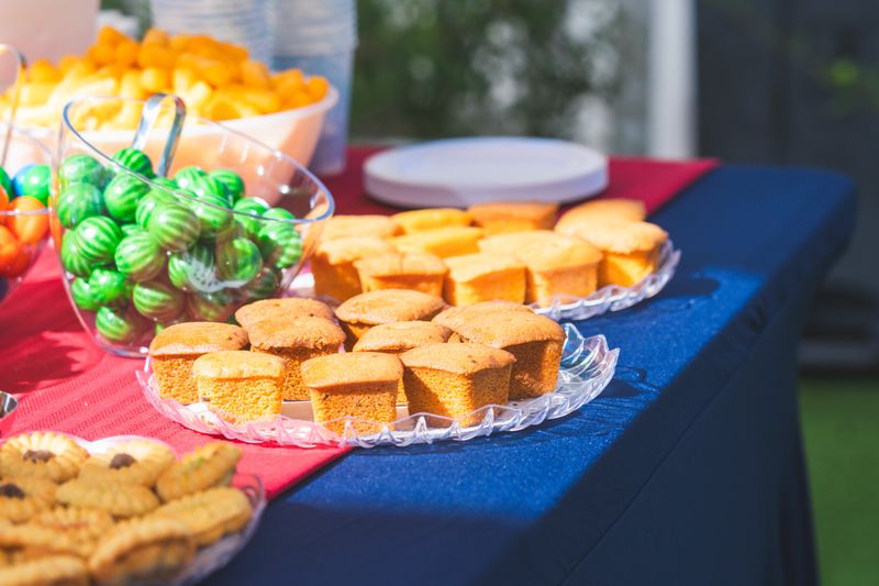 Assorted bite-size cakes, cream cookies, mini cupcakes, and a jar of colorful candy arranged on a decorated outdoor party table, creating a festive and inviting dessert spread in natural daylight.