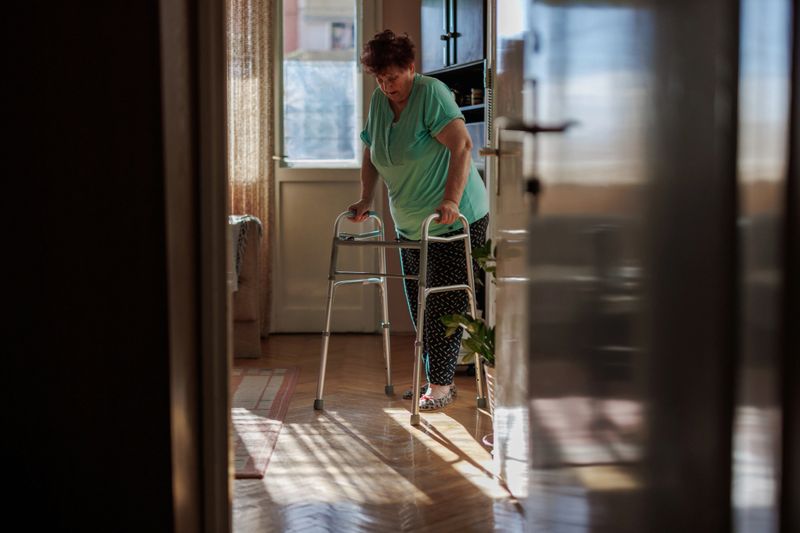 An older woman uses a walker for support while moving through her living room , illuminated by natural light from a window