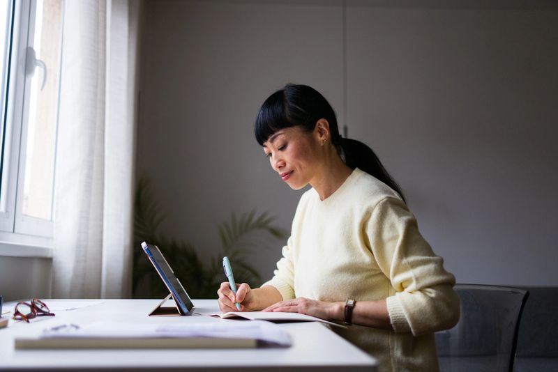 A calm, focused woman writes in a notebook at a bright desk, with a tablet nearby.