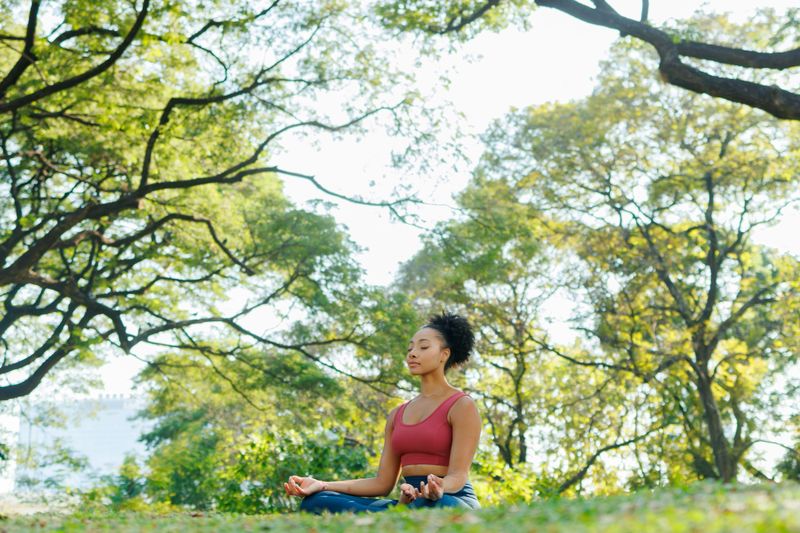 A serene athletic woman practicing mindfulness and spiritual wellness sitting in lotus pose meditation, resting her hands in Gyan Mudra on her knees while sitting on a yoga mat on the green grass of a peaceful park in the morning.