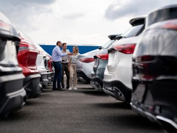Three people discussing between rows of parked cars in a lot.