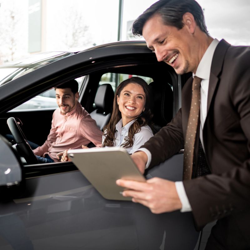 Happy couple engaging with a car salesman at a dealership, discussing vehicle options and purchase terms on a digital tablet