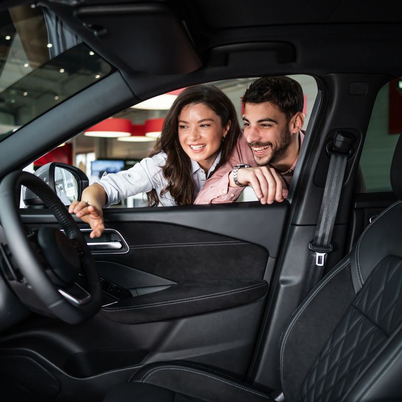 Young couple smiling, looking at a new car interior. They are shopping for a vehicle inside a modern auto showroom