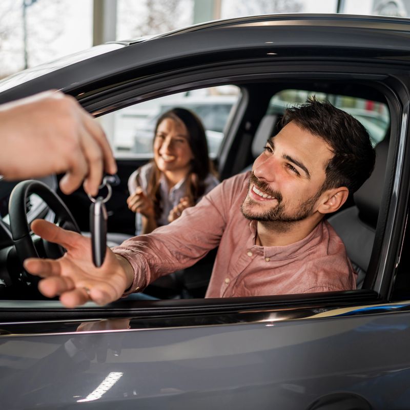Smiling man reaching for new car keys from salesperson while woman sitting inside vehicle feeling happy