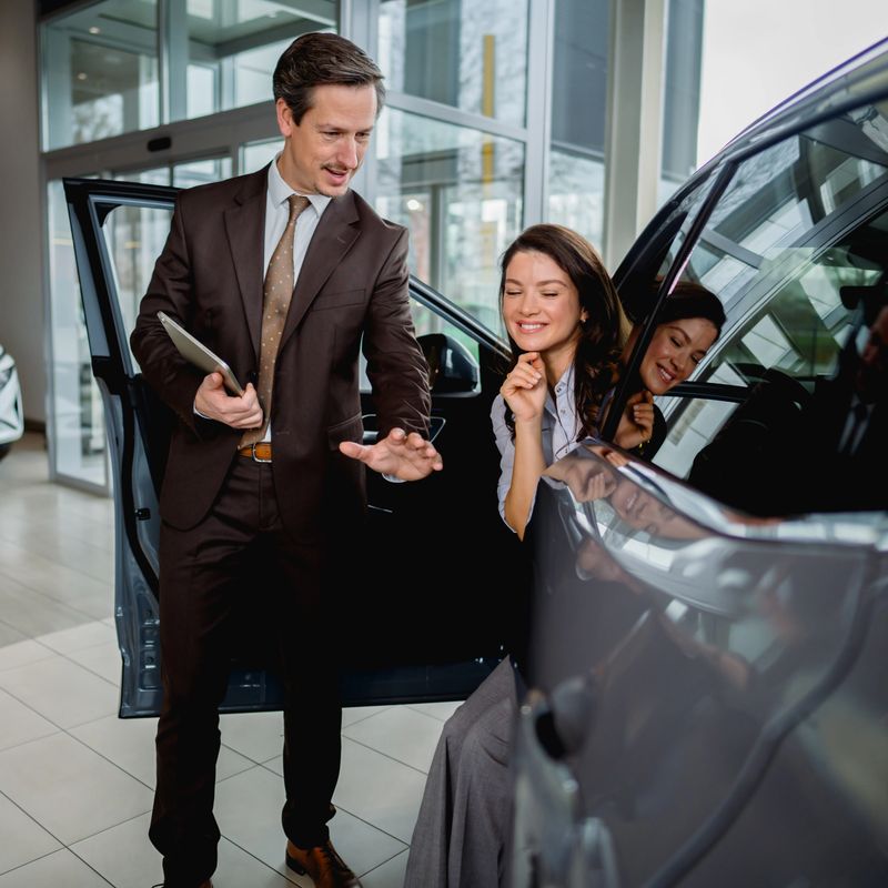 Car salesman presenting features of a new vehicle to a smiling woman sitting inside at an automotive dealership