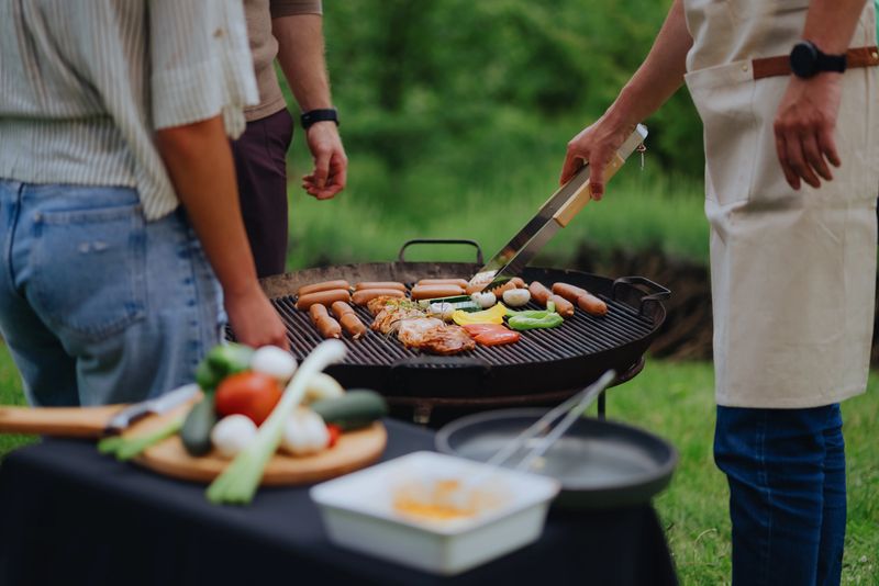 People enjoying a summer barbecue, grilling sausages, chicken, and vegetables on a hot grill in a relaxed outdoor setting