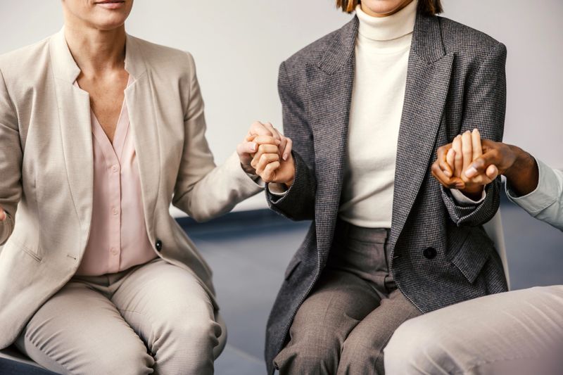 Diverse group of adults sitting close, holding hands in a supportive circle indoors, expressing unity, trust and empathy during group therapy or recovery meeting