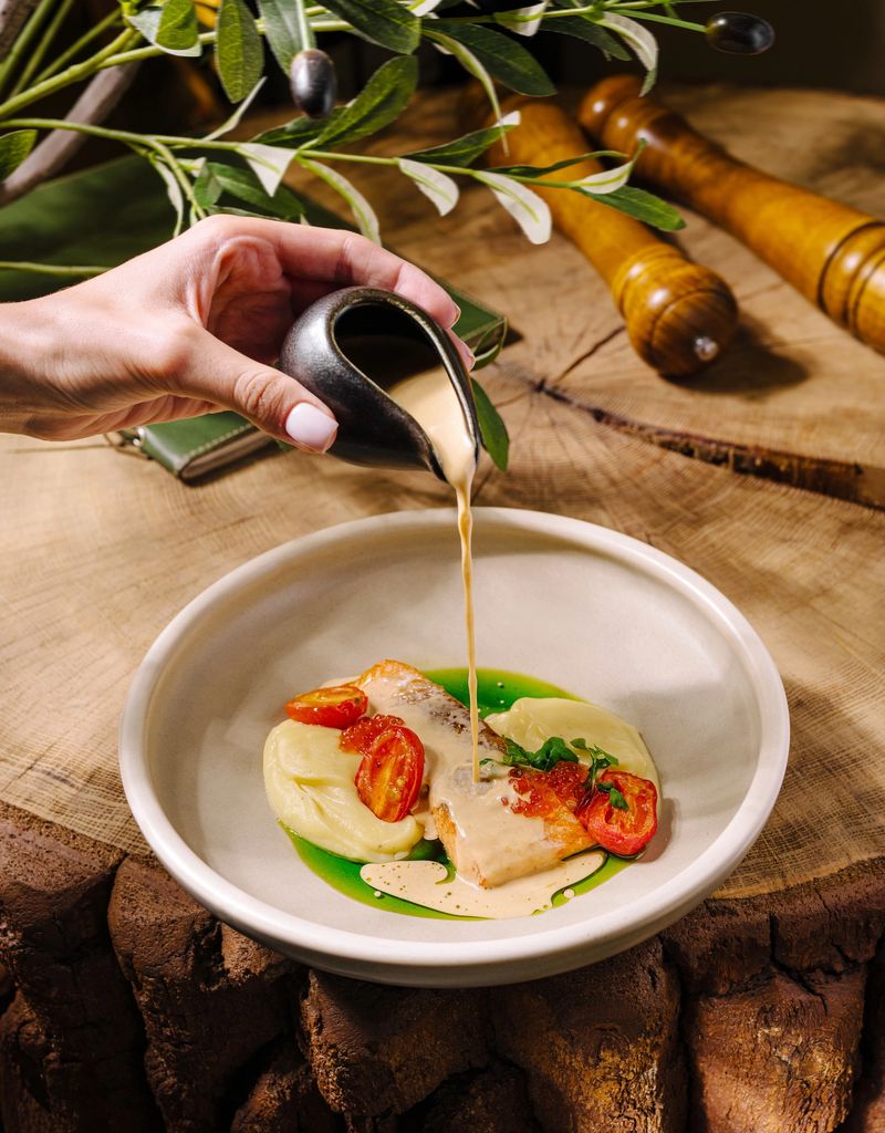 A hand elegantly drizzles sauce onto a plate with salmon, tomatoes, and creamy puree. The rustic setting includes wooden elements and a touch of greenery.