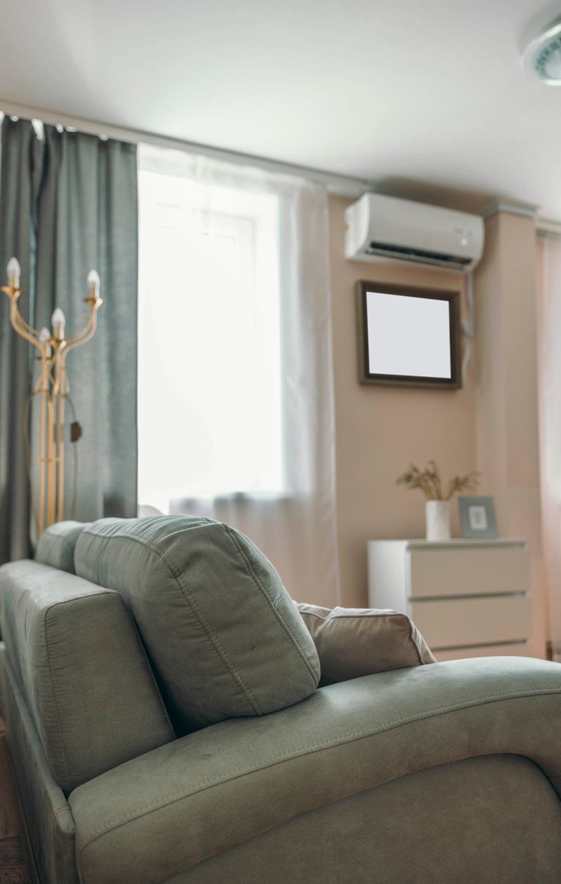 Cozy hotel room interior with a comfortable gray sofa, air conditioning unit, and natural light from a window. Minimalist decor with a small table and artwork.