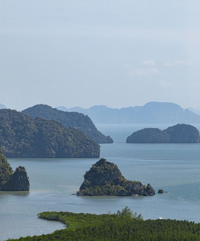 Panoramic tropical seascape with dramatic limestone karst islands emerging from calm turquoise water, surrounded by lush green mangrove forest and distant island silhouettes under a blue sky. Iconic coastal landscape showcasing untouched nature, geological formations, and serene atmosphere. Ideal for travel, tourism, adventure, conservation, environment, and exotic destination concepts. No people.