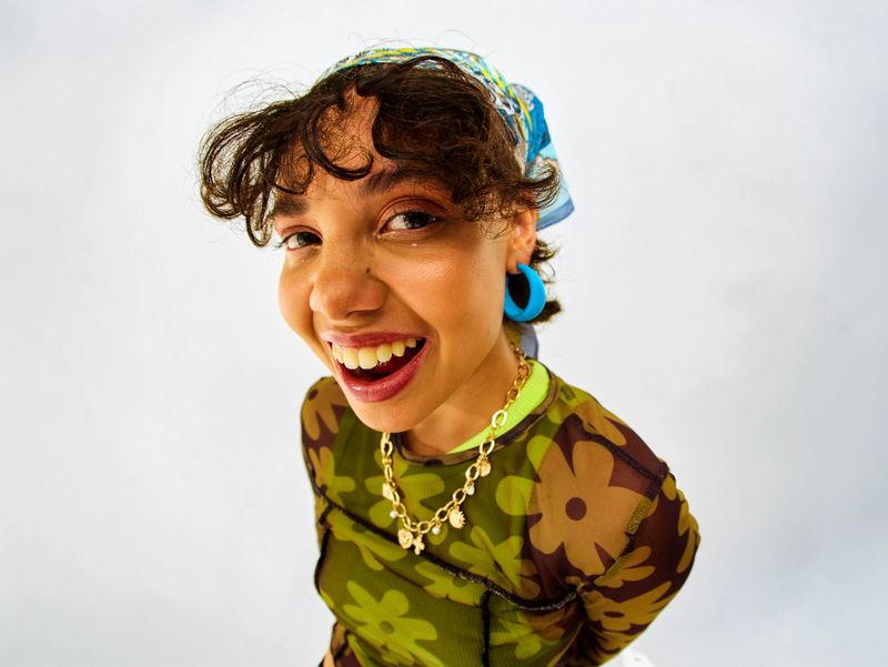 Young adult woman wearing floral mesh top, gold chain, and blue earrings in studio