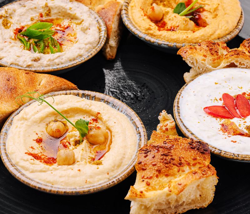 A vibrant display of four types of hummus accompanied by pieces of warm bread. The spread includes chickpeas and garnishes, perfect for sharing at any gathering.