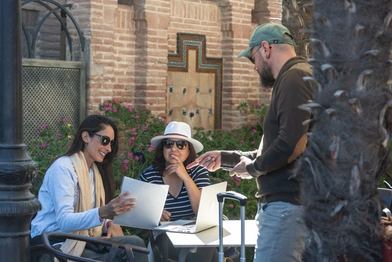 Three friends having an informal business discussion at an outdoor cafe, reviewing documents and using a laptop, enjoying a relaxed meeting