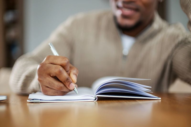 Young black man concentrating while writing notes in a personal journal or notebook with a pen, capturing thoughts and ideas for personal development and creative expression