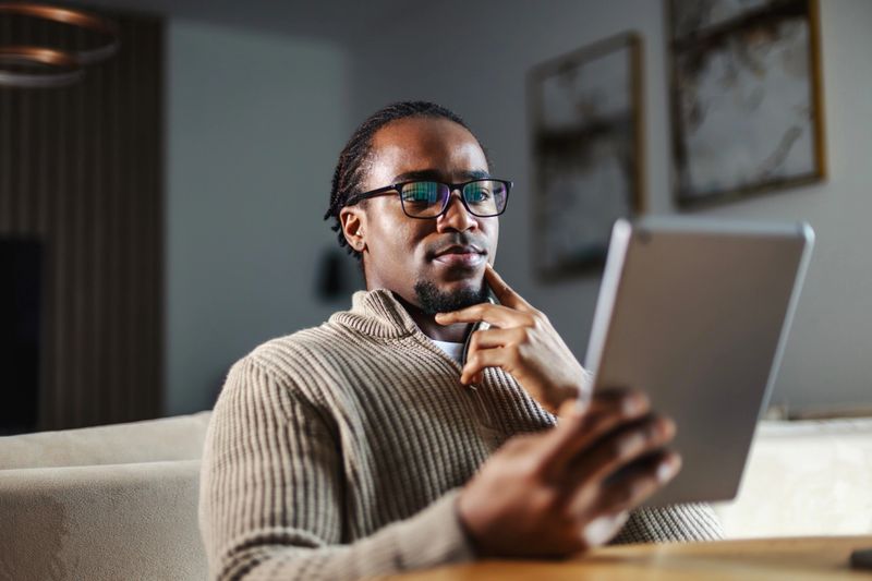 Thoughtful african american man with eyeglasses sitting at a table, focusing intensely on his digital tablet while learning or working remotely from home